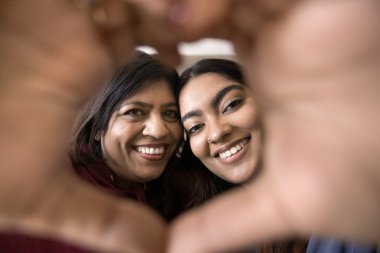 İki mutlu Hintli kadın, aile üyeleri, kameraya el yapımı kalp şeklinde bakın, beyaz dişleri gösterin, dişçi reklamı yapın. Anne ve kızı arasındaki koşulsuz sevgi. Kapatın.