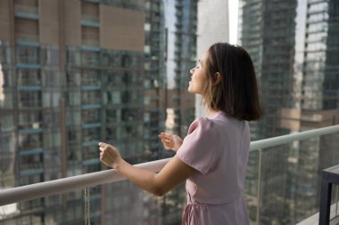Rahat kadın, modern balkonda, gökdelenlere tepeden bakan şehir manzarasında duruyor, gözleri kapalı sükunetin tadını çıkarıyor, huzur, özgürlük hissi yayıyor, kentsel yaşamla bağlantı kuruyor.