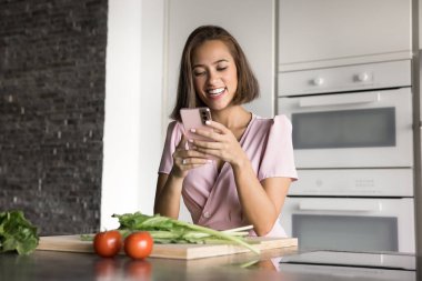 Akıllı telefonunu kullanan güzel bir kadın rahat bir mutfakta sohbet eder, yemek tarifi okur, mobil mutfak uygulamaları indirir, çevrimiçi süpermarket aracılığıyla sipariş verir. Aşçılık ve modern teknoloji