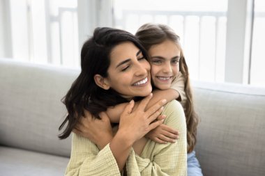 Küçük kız, genç ve güzel annesine sıkıca sarılıyor, koşulsuz yaşadığını hissediyor, şefkatini ifade ediyor, oturma odasında kanepede oturuyor. Anne-kız bağı, yakınlık. Portre, aile anları