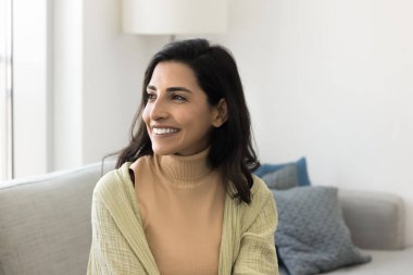 Brezilya 'lı güzel bir kadın günlük giysiler içinde oturma odasında rahat bir koltukta tek başına oturuyor sıcak bir gülümsemeyle, neşeli bir ifadeyle mesafeyi seyrediyor. Dinlen, evde kaygısız vakit geçir.