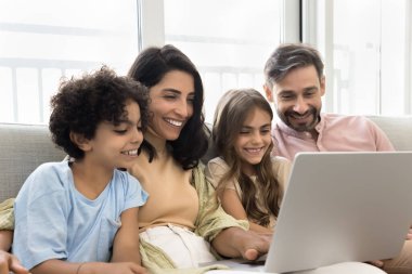 Çocuklu bir çift kanepeye oturup dizüstü bilgisayarla video izliyor, aile fotoğraflarına bakıyor, arkadaşlarıyla veya akrabalarıyla görüntülü görüşme yapıyor, gülümsüyor, neşe dolu mutlu aile anlarının tadını çıkarıyor.