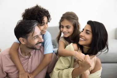 Dört çocuklu neşeli aile, çoklu etnik çeşitliliğe sahip genç aileler ve küçük çocuklar evdeki kanepede oturmuş birbirlerini kucaklıyorlar, aile sıcaklığı ve mutluluk yayıyorlar, sevgiyi hissediyorlar. Bond ve eğlence