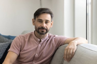 Orta yaşlı, kısa saçlı ve sakallı Portekizli bir adam, günlük pembe gömlek giyiyor, oturma odasında koltukta oturuyor, ciddi bir ifadeyle kameraya poz veriyor, rahat ve sakin görünüyor, ev portresi yapıyor.