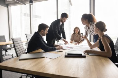 Stratejik proje değerlendirme toplantısı. Çok ırklı iş adamlarından oluşan bir grup, pazarlama uzmanları brifinge katılıyor, yaklaşan projeyle ilgili belgeleri gözden geçiriyor, fikirleri paylaşıyor, araştırıyor, tahmin yürütüyor