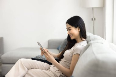 Yan manzara güzel Asyalı kadın cep telefonuyla koltukta oturuyor, mesajlaşmanın keyfini çıkarıyor, kurye başvurusu yoluyla, haberleri okuyarak, sosyal medyada kayarak, internette yalnız başına vakit geçiriyor.