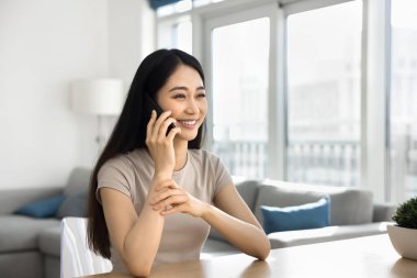 Güzel Asyalı kadın rahat oturma odasında masada oturuyor, gülümsüyor, akıllı telefonda hoş sohbetlere öncülük ediyor, iyi haberler alıyor, telefonla olumlu iletişimin tadını çıkarıyor. Modern teknoloji