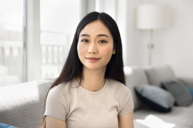 Güzel Asyalı bir kadının portresi. Rahat, güneşli, aydınlık bir odada oturuyor. Huzurlu bir ifadeyle kameraya bakıyor. Güven, modern yaşam tarzı, ev konforu, kişisel refah.