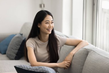 Güzel, dingin Çinli bir kadın rahat bir kanepeye oturmuş, sıcak bir gülümseme ile pencereden dışarı bakıyor, hoş düşüncelerin derinliklerinde, hafta sonu evde tek başına dinlenirken yansıtıyor. Hayal kurmak, rahatlamak.