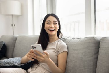 Asyalı kadın gülüyor, modern akıllı telefonu evde kullanıyor, başka tarafa bakıyor, hoş online haberlerin tadını çıkarıyor ya da erkek arkadaşla sohbet ediyor, uyarı alıyor, internetin içinde kaygısızca eğleniyor. Teknoloji, eğlence.