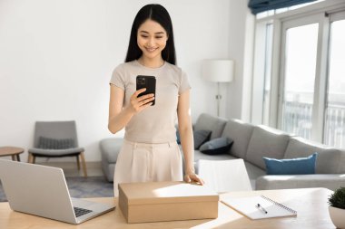 Tatmin olmuş genç Asyalı kadın rahat oturma odasında cep telefonuyla, paket kutusuyla masada duruyor, gülümseme mobil uygulama aracılığıyla sipariş edilen kurye servisinin keyfini çıkarıyor, olumlu geri bildirim yazıyor.