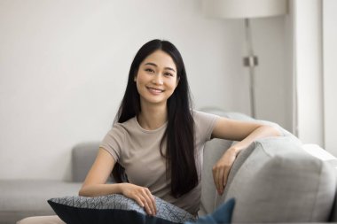 Ev portresi için poz veren genç Asyalı bir kadın, oturma odasında rahat bir şekilde oturuyordu, modern iç mekanı olan, dişlek gülüşlü kameraya bakıyordu. Güzel kadın model, profil resmi.