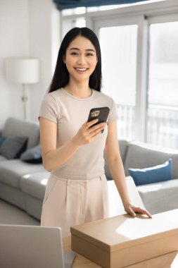 Asyalı internet mağazası müşterisi, oturma odasında akıllı telefonla poz veriyor, karton kutu alıyor, ulaşım lojistik hizmetleri ya da teslimat paketi için e-ticaret uygulaması kullanıyor,