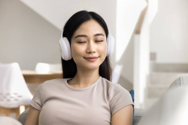 Huzurlu Çinli kadın kanepeye oturmuş, gözleri kapalı, modern kablosuz kulaklık takmış, evde tek başına dinlenen favori şarkıyı seviyor. Dijital akış aboneliği müzik hizmetleri uygulaması kullanıcısı