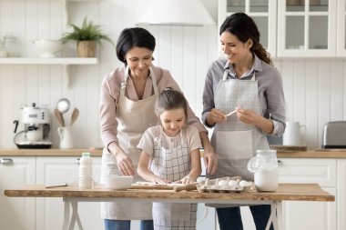 Komik aile yemekleri. Mutlu üç kadın nesil olgun büyükanne küçük kız önlüklü küçük kız tatlı pişirmek için mutfak masasında toplanıyor ev yapımı fırın için hamur yapıyor.