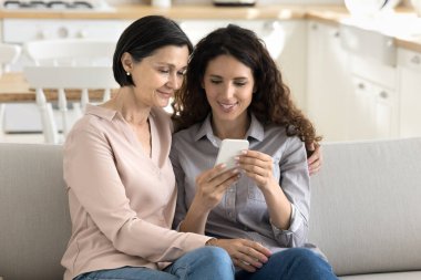 Modern teknoloji ve ortak ilgi alanları. Mutlu aile anları. Yetişkin anne kızı koltukta sarılıyor. İnternetten alışveriş videolarını izliyorlar. Boşluğu kopyala