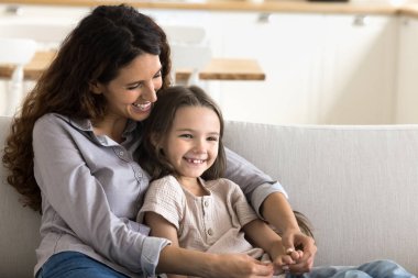 Gerçek aşk. Mutlu gülümseyen genç anne, sevimli küçük kızını sıkıca kucağına alıp sıcak kanepeye oturur sıcak şefkatli çocukların avuçlarını okşar ve çocuğuyla huzurlu bir sohbetin tadını çıkarır. Boşluğu kopyala
