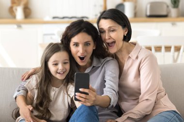 İnanabiliyor musun? Dost canlısı bir aile büyük anne küçük kız kanepede sarılıyor heyecandan çığlık çığlığa telefon ekranına bak piyango ödülünü kazan ecommerce mağazasından inanılmaz bir sürpriz geliyor