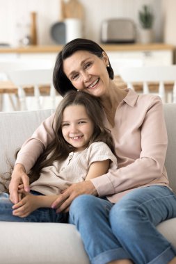 Büyükannenin sevgi dolu kucaklamalarıyla. Dikey fotoğraf portresi sevimli küçük kız rahat koltukta oturuyor sevgili büyükannesine yaslanıyor kameraya bakıyor kendini güvende hissediyor ilgili büyükannelerin kollarında.