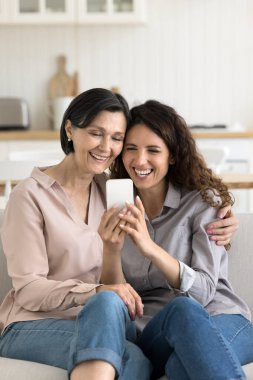 Aile eğlence zamanı ve dijital teknoloji. Samimi gülüyor genç kız yaşlı anne mutfakta kanepede sarılıyor rahatla cep telefonundaki komik selfie fotoğrafını çek komik videoların keyfini çıkar komik mesajları oku