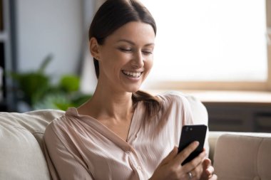 Güzel, gülümseyen bir kadın elinde akıllı bir telefonla oturmuş, hoş mesajlar okuyor, erkek arkadaşından mesajlar alıyor, uzaktan iletişim kuruyor, uygulamaları kullanıyor, eğleniyor, çevrimiçi eğlence kavramını kullanıyor.
