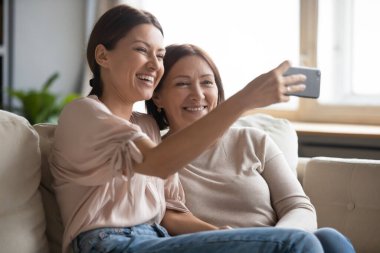 Güzel, yetişkin bir kız ve orta yaşlı bir anne kanepede oturmuş fotoğraf çekiyor. Evde eğleniyor, fotoğraf çekiyor, fotoğraf çektiriyorlar. Akıllı telefonlu selfie çekiliyor.