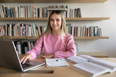 Genç 20 'li yaşlarda neşeli bir öğrenci bilgisayar ve çalışma materyalleriyle dolu masaya oturmuş kameraya bakar, konu öğrenirken veya kütüphanede sınava hazırlanırken kafasında doğru duruş için kitap dengesi kurar