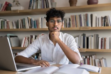 Üniversite kütüphanesinde kameraya poz veren ciddi Hintli öğrencinin portresi, egzersiz yapmak, özet yazmak, dizüstü bilgisayarla ders kitaplarını kullanarak tek başına çalışmak. Yüksek öğrenim, kalkınma