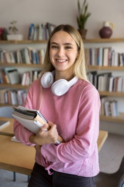 Kütüphanede ya da kolej sınıfında ders kitapları ve çalışma kitaplarıyla tek başına duran gülümseyen kız fotoğrafına bakın. Kameraya bakın. Mükemmel öğrenci portresi. Üniversite giriş ve yükseköğretim