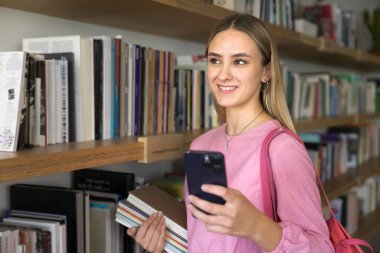 Kütüphanede dururken akıllı telefon kullanan, çalışma kitaplarını tutan, mesajları paylaşan, modern mobil uygulama programı kullanarak edebiyat veya okul sınıflarını kontrol eden düşünceli bir kız öğrenci.
