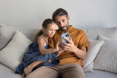 Olgun Portekizli adam, küçük kızı akıllı telefon, mesajlaşma, video arama, yeni yapay zeka mobil uygulamalarını keşfetme, video oyunu oynama veya pazar platformu üzerinden hizmet satın alma gibi konularda kanepede dinleniyor.