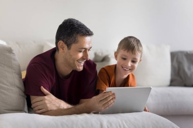 Baba ve küçük oğlu kanepede tabletle dinleniyorlar, video izliyorlar, eğitim içerikleri var, bulmaca oynuyorlar, interaktif öğrenmenin tadını çıkarıyorlar, dijital hikaye kitapları okuyorlar, boş zamanlarını çizim yaparak geçiriyorlar ve uygulamalar yaratıyorlar.