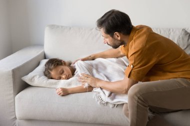 Orta yaşlı baba sıcak ekoseyle üstünü örtüyor. 6 'lı yaşlarındaki küçük kızı oturma odasında rahat bir kanepede uyuyor. Gündüz uykusunda rahat ve iyi bir uyku çekiyor. Ebeveyn sevgisi, çocuk bakımı, rahatlama.