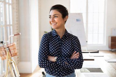 Genç Hintli iş kadını modern yaratıcı ofiste poz veriyor, gülümsüyor, başka tarafa bakıyor, gelecekteki mesleki başarılar hakkında derin düşünceler besliyor, tamamlanmış projelerden tatmin oluyor, kurumsal hedeflere ulaşıyor.