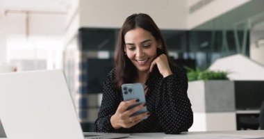 Çevrimiçi içeriği iş yerinde akıllı telefondan izleyen, ekommerce uygulamasını kullanan, iş iletişimi için internet hizmeti veren, gülümseyen güzel Orta Doğulu iş kadını.