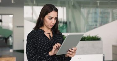 Ciddi şekilde odaklanmış Ortadoğulu profesyonel iş kadını internet hizmetini kullanıyor, iş için tablet üzerinde internet iletişimi kuruyor, ofis yerinde aletlerle duruyor, mutlu oluyor, gülüyor.