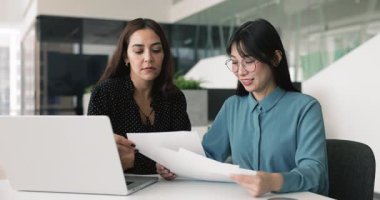 Kağıt pazarlama raporunu analiz eden, belgelerde mali istatistik verilerini birlikte inceleyen, ürün yönetimi ve strateji tartışan iki farklı kadın çalışan.