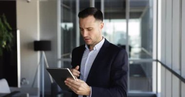 Ciddi düşünceli Latin iş adamı, tablet üzerinde finansal internet hizmetini kullanıyor, ödeme yapıyor, işlem yapıyor, yatırımları analiz ediyor, ofisinde daktilo yazıyor, başka yere bakıyor, çeneye dokunuyor.