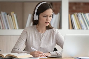 Modern Bluetooth kulaklık takan odaklanmış milenyum kızı dizüstü bilgisayara bak, not al, kulaklık takan konsantre kız öğrenci bilgisayarı kullanarak sınava hazırlanıyor.
