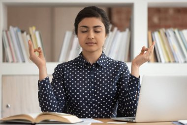 Sakin Hintli kız öğrenci masasında oturmuş Mudra elleriyle arabuluculuk yapıyor, ders çalışmaya ara veriyor, rahatlamış milenyum kızı iş yerinde yoga yapıyor, temiz hava soluyor, stresten uzak konsept yapıyor.
