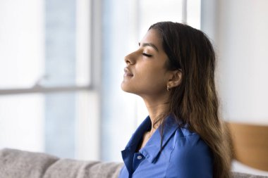 20 'li yaşlardaki genç Hintli kadın kanepeye oturmuş, gözleri kapalı, temiz hava soluyor, derin bir rahatlama veya meditasyon anında, yüzünü kapatıyor. İç huzur, stres atma, nefes alma teknikleri