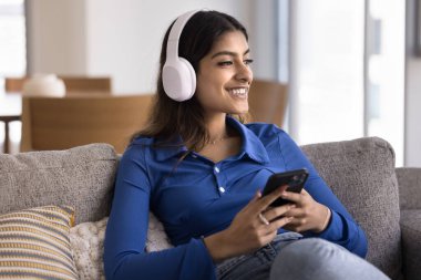 Hintli bir kadın koltukta rahatça oturuyor, kulaklık takıyor, telefonu tutuyor, favori müzik ve podcast 'ten hoşlanıyor. Modern teknoloji, çevrimiçi dijital abonelik hizmetleri platformu kullanıcısı yoluyla rahatlama