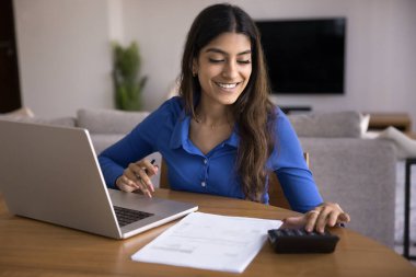 Kadın dizüstü bilgisayarla, hesap makinesiyle ve evrak işleriyle masada oturuyor, banka hesaplarını kontrol ediyor, mali raporlar hazırlıyor, vergi beyannameleri ya da mali belgeler üzerinde çalışıyor. yaklaşan ödemeleri ya da giderleri planlamak