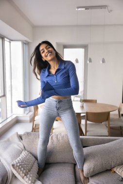 Dikey mutlu Hintli kadın evdeki kanepede dans ediyor, gülüyor, tatmin oluyor, hayatı kutluyor ya da ısınıyor, tek başına sorunsuz bir hafta sonu tatili geçiriyor ya da ev kiralıyor.