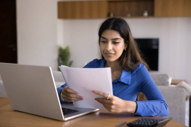 Genç Hintli kadın dizüstü bilgisayarında kağıt tutarak masada oturuyor, evrakları inceliyor, inceliyor, analiz ediyor, işle, finansla ya da projeyle ilgili, anlaşma şartlarını ve şartlarını okuyor, kağıtları düzenliyor.