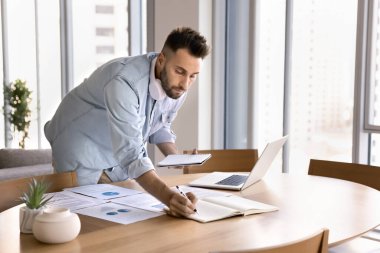 Odaklanmış adam çoklu görev, analitik görev üzerinde çalışmak, sunum hazırlamak veya masada duran araştırmaları yönetmek, yazı, dizüstü bilgisayar, yazılı belgeler çalışma masasına dağıtılırken telif defteri tutmak