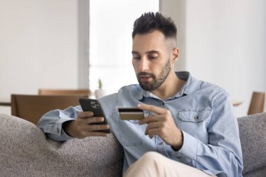 Odaklanmış İspanyol bir adam evinde oturmuş banka kartı ve akıllı telefon kullanarak online alışveriş yapıyor, e-ticaret platformuna bakıyor, mal sipariş ediyor, güvenli e-banka uygulaması üzerinden çevrimiçi ödemeler yapıyor.