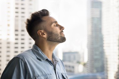 Yansıtıcı, sakin İspanyol bir adamın derin, sakin bir nefes alıp, gözleri kapalı huzur anının tadını çıkardığı, büyük şehir manzarasına karşı balkonda durduğu profil yüzü. Farkındalık, hayaller