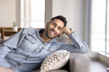 Genç Latin bir adam rahat, modern oturma odasında rahat bir koltukta dinleniyor, çağdaş konutta tek başına vakit geçiriyor, neşeli ve memnun bir ifadeyle kameraya bakıyor. Evde rahatlama