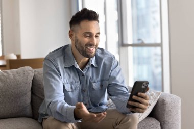 Latin adam rahat bir şekilde koltukta oturmuş elinde akıllı telefonuyla canlı bir video görüşmesine bağlanıyor cep telefonu uygulaması kullanıyor, gülümsüyor, hoş haberler alıyor, güncellemeler alıyor, yeni yazılımın tadını çıkarıyor.